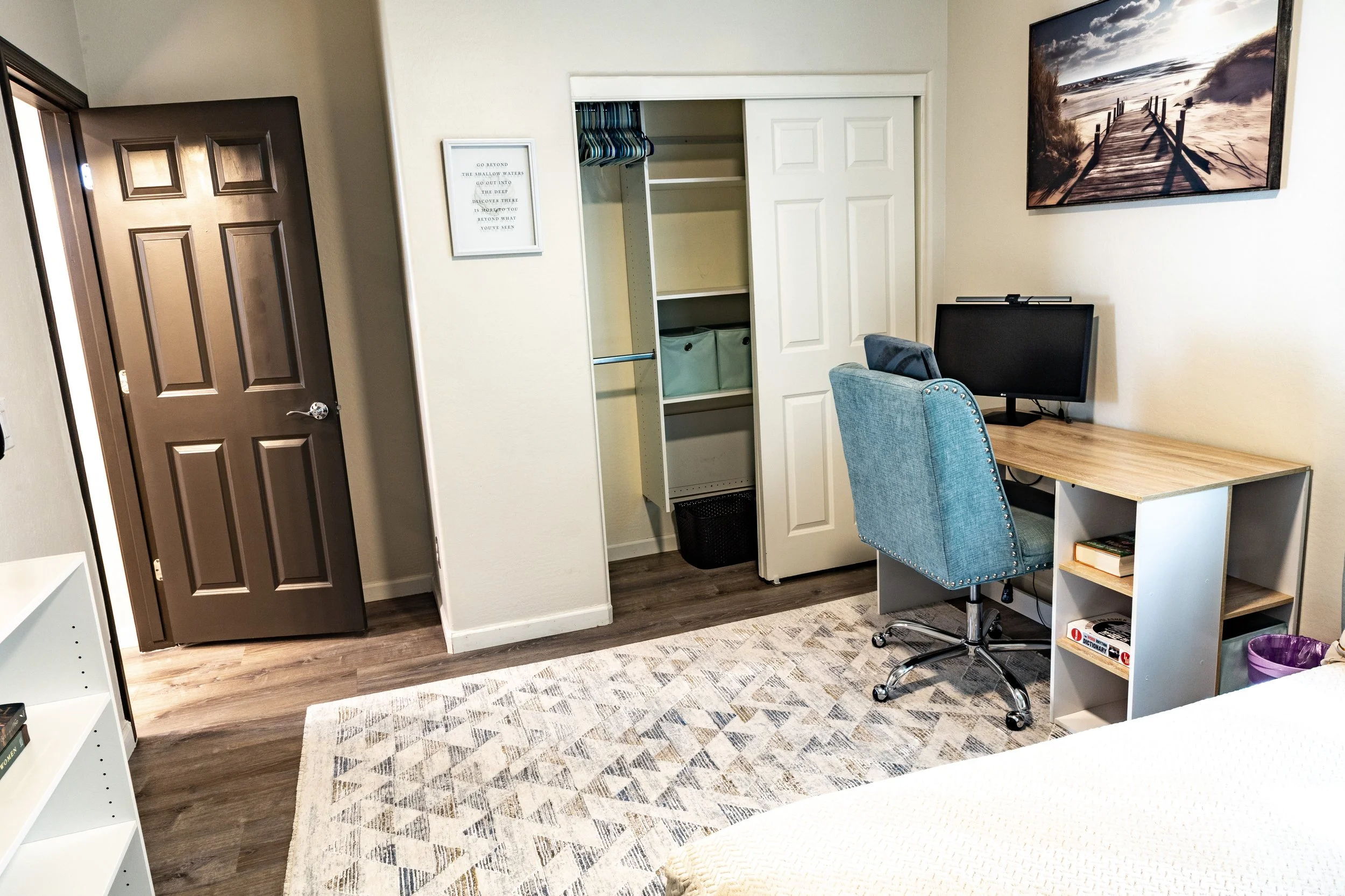A cozy home office with a wooden desk, a swivel chair, a desktop monitor, and a bookshelf. There is a closet with sliding doors, and a large landscape painting on the wall depicting a beach scene with a pier. The floor has dark wood laminate, and a p