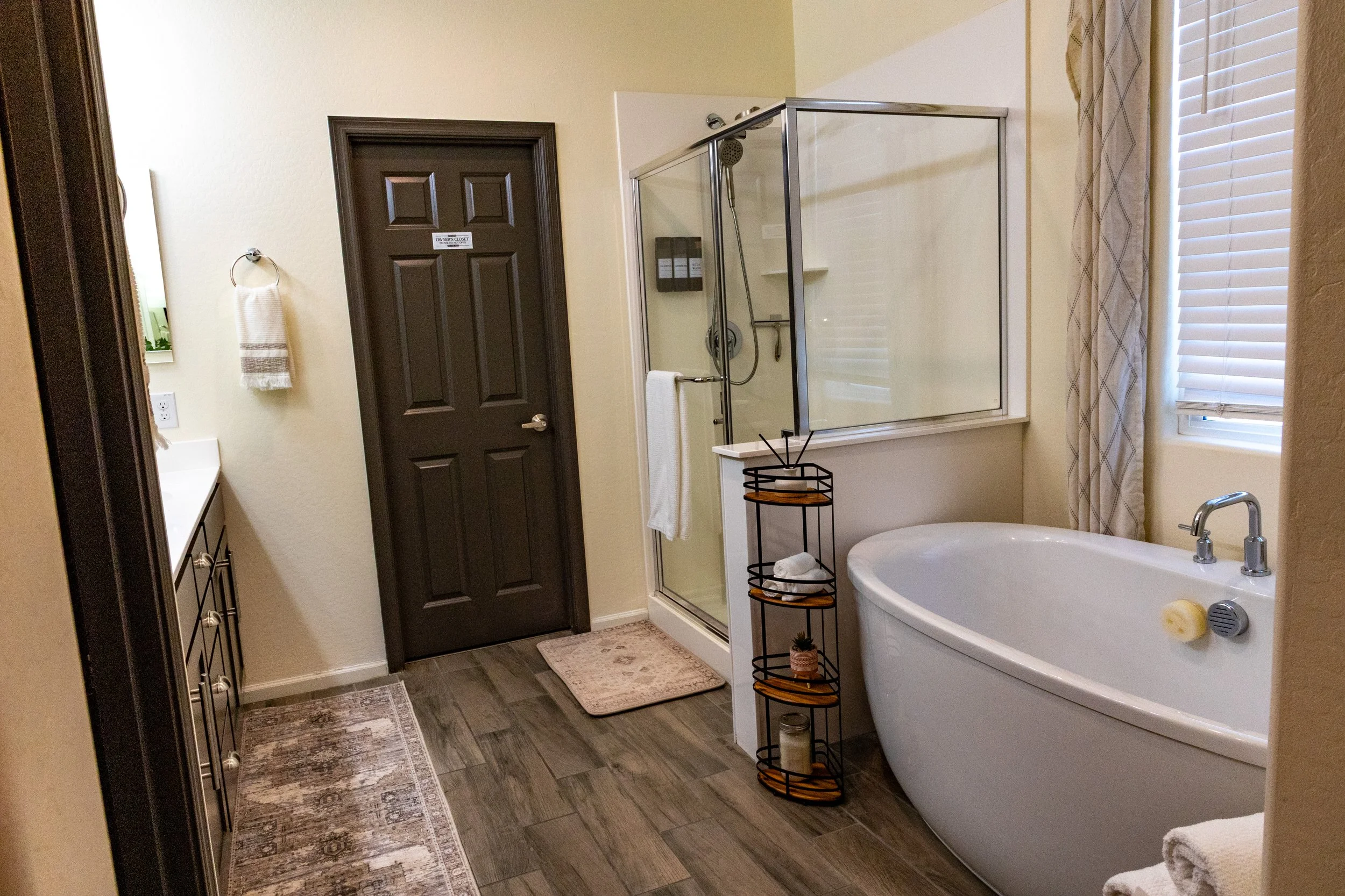 A bathroom with a dark brown door, a glass shower enclosure, a bathtub, and a window with curtains. There are towels hanging on towel rings and a small black metal rack with shelves next to the bathtub, holding towels and small decorative items. The 