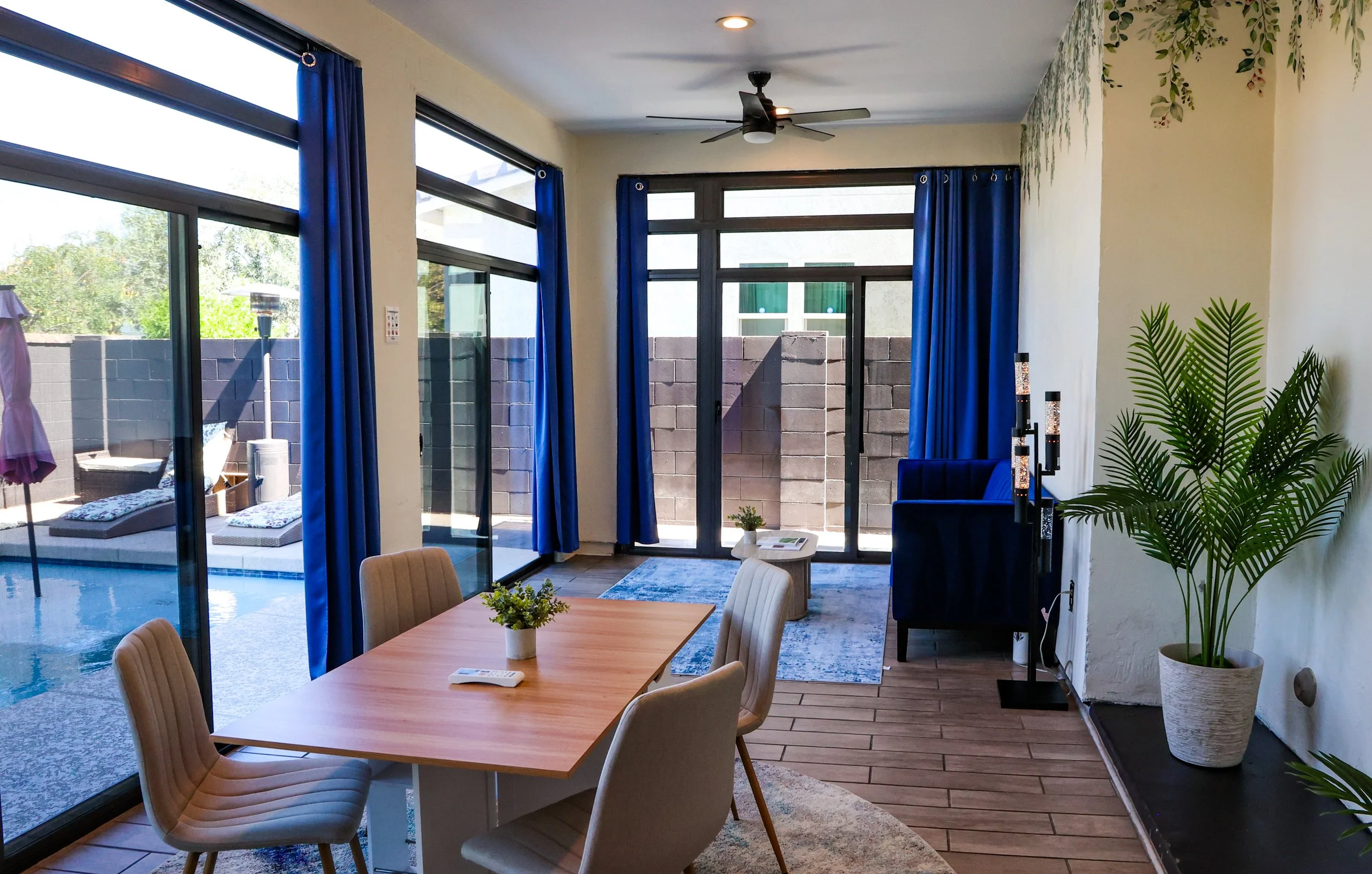 Living room with large windows, blue curtains, ceiling fan, and a view of an outdoor seating area with a pool. Decor includes a blue armchair, plants, and a small table.