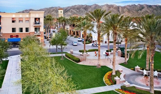 A landscaped urban area with palm trees, a green lawn, a walkway, and a stone staircase. Surrounding buildings and a mountain range in the background.