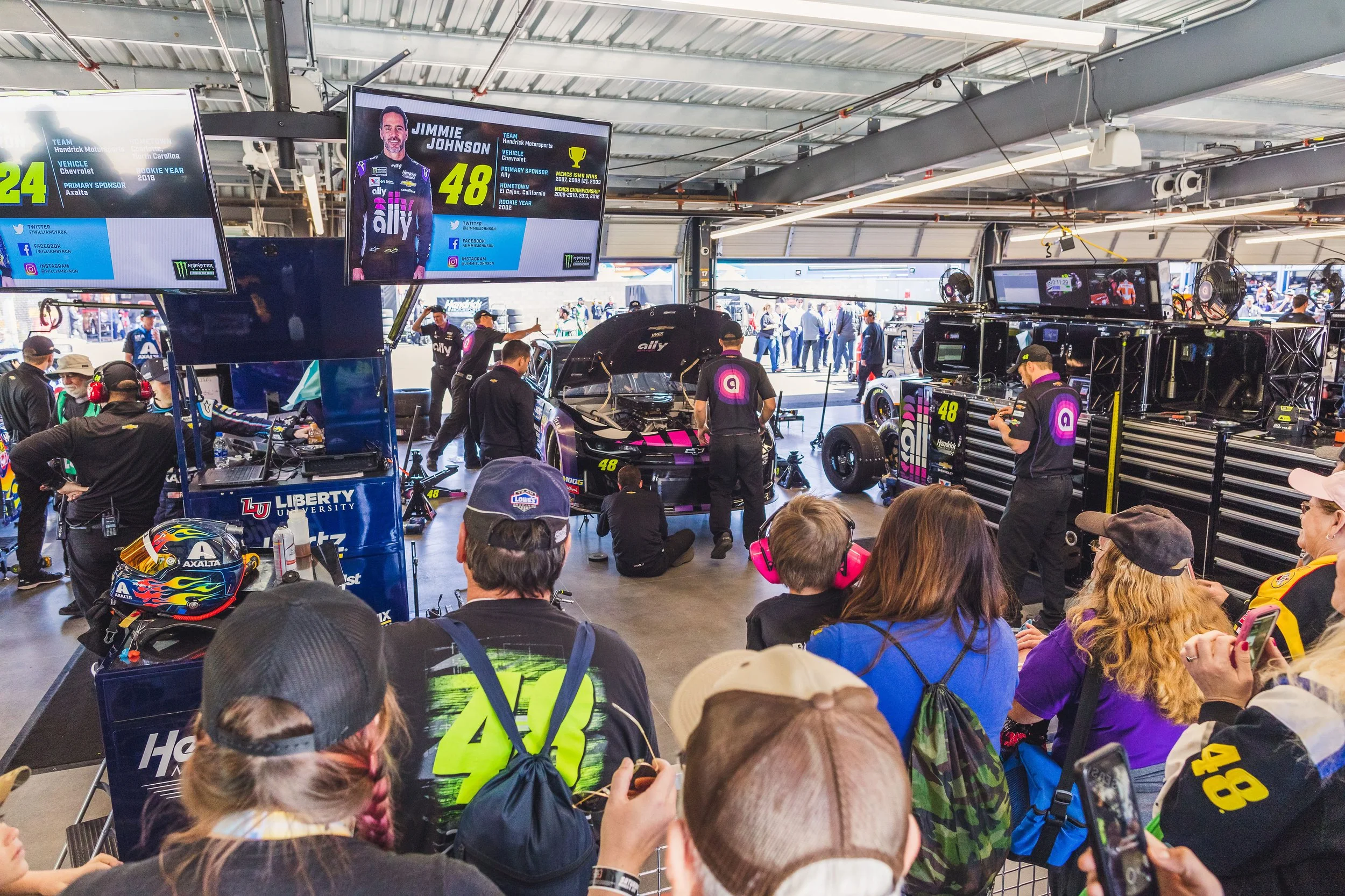 The image shows a busy garage area at a racing event with multiple people, race cars, and equipment. A large screen displays a driver profile, Jimmie Johnson, with team and sponsor information. Fans and crew members are engaged in various activities 