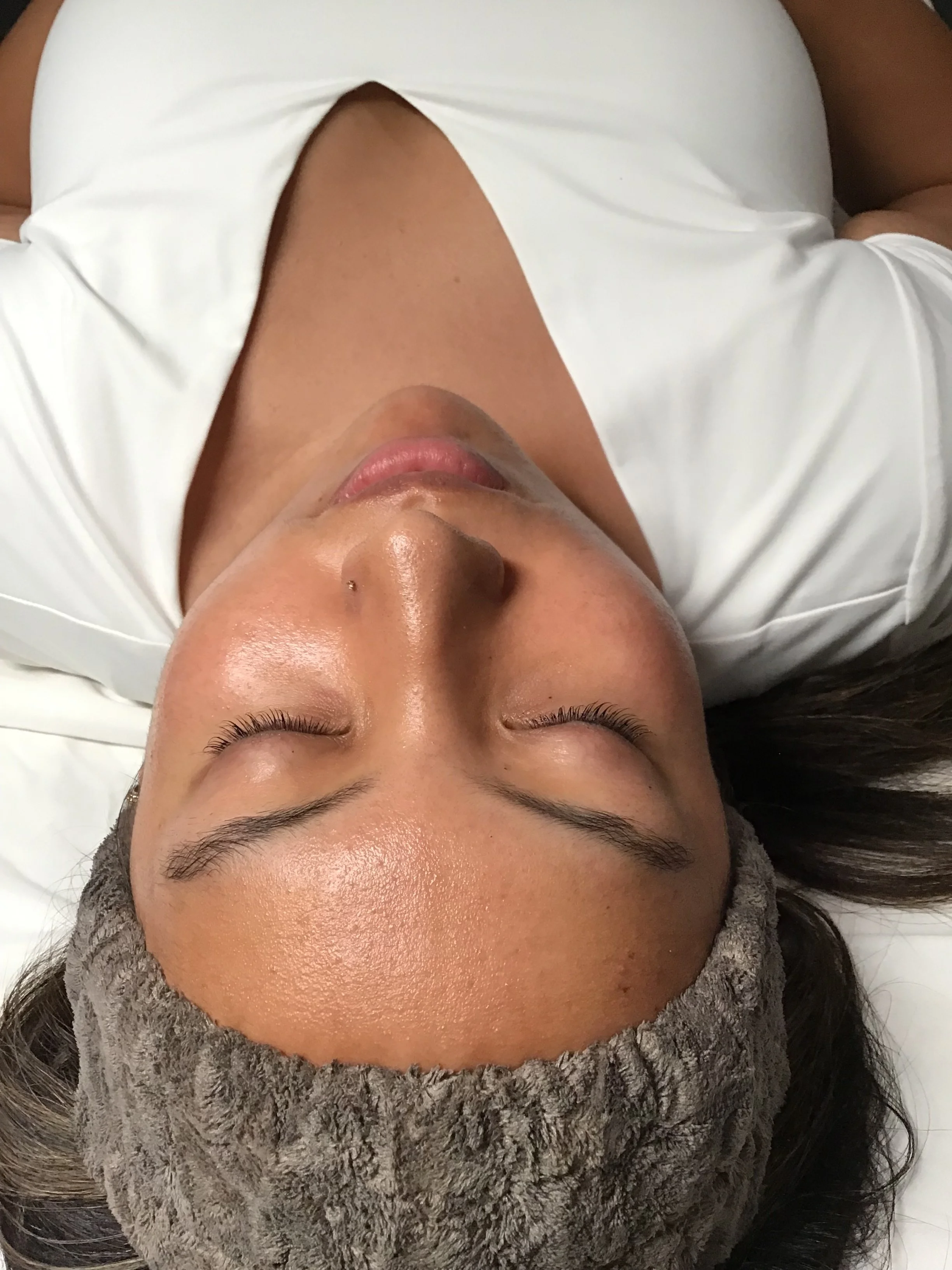 A woman lying on her back with eyes closed, wearing a white top and a gray headband, showing her face, forehead, and closed eyes.