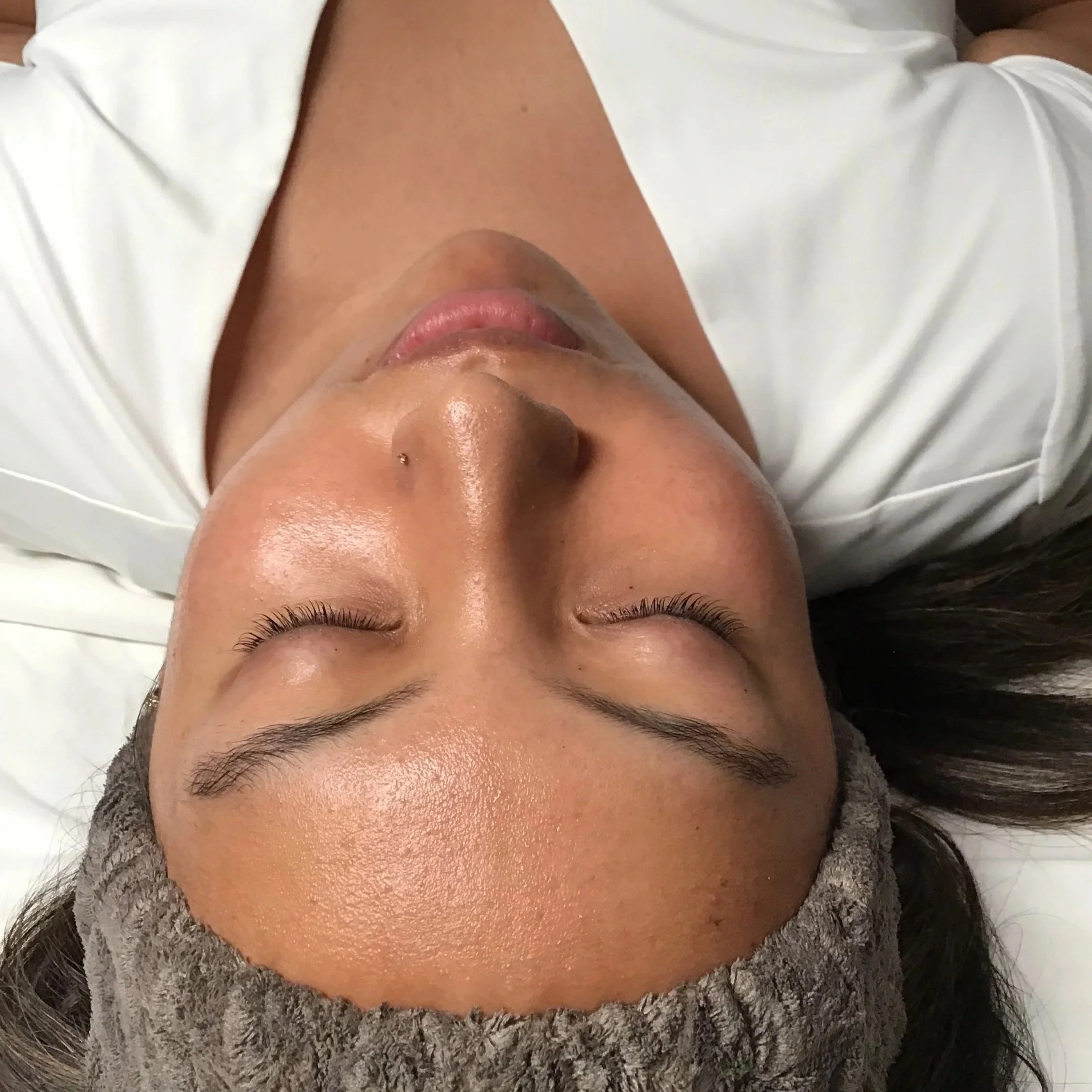 Close-up of a person's face lying down with closed eyes, wearing a white garment and a gray textured scarf, showing smooth skin, arched eyebrows, long eyelashes, and a small nose piercing.