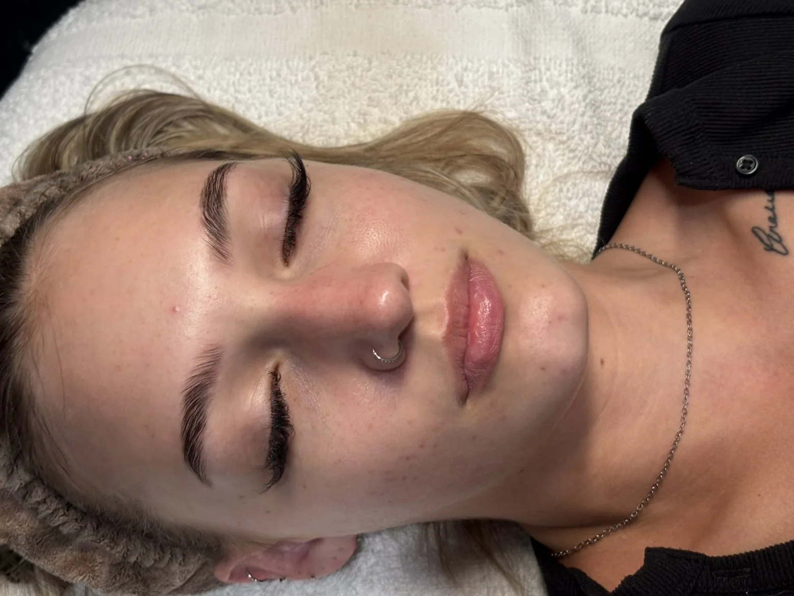 A woman with closed eyes lying down, with a nose ring, wearing a black shirt, a chain necklace, and a knit headband.