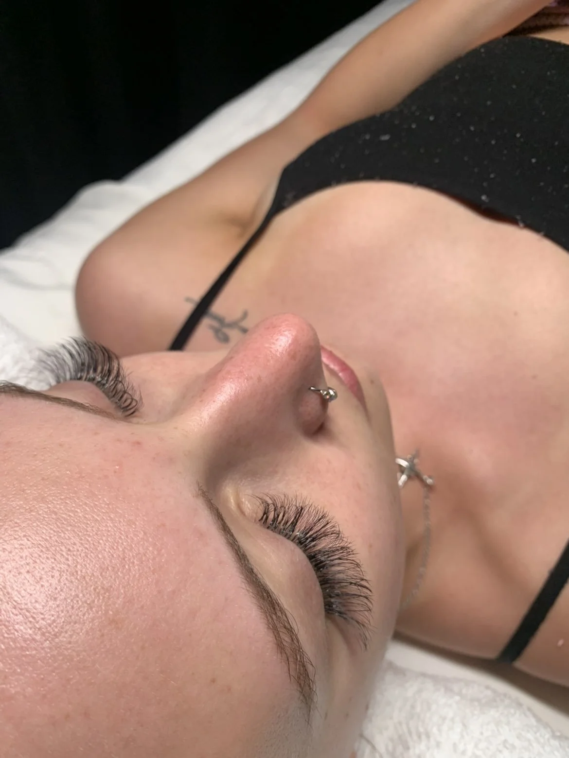 A woman with long eyelashes, piercing on her nose, and a tattoo on her shoulder, lying down on a bed.