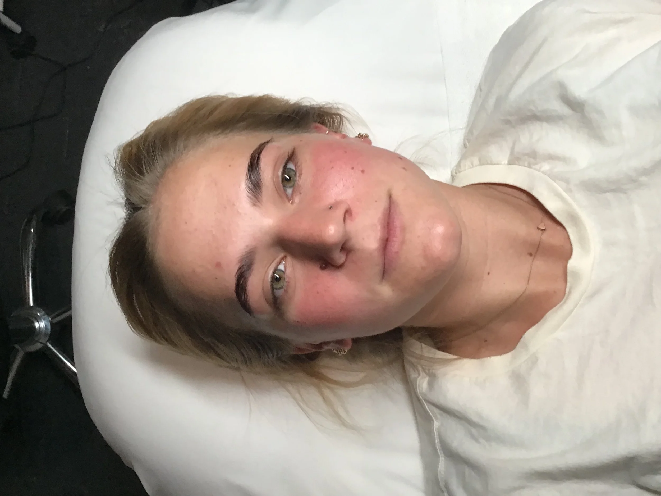 A woman with light brown hair and tattoos resting on a hospital bed with white linens, looking at the camera.