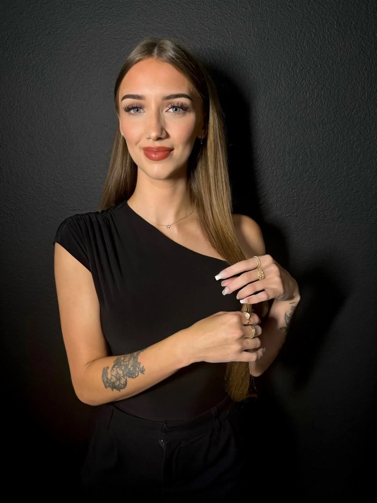 Portrait of a young woman with long brown hair, wearing a black top, standing against a black wall, smiling softly and holding her hands near her chest.