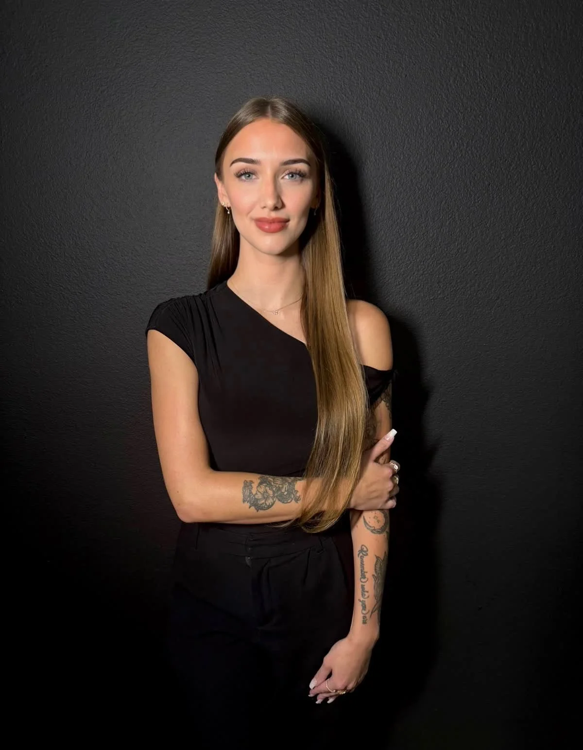 A young woman with long straight hair wearing a black top with one shoulder cut out, standing against a black textured wall, smiling softly with her arms crossed and tattoos visible on her forearms.