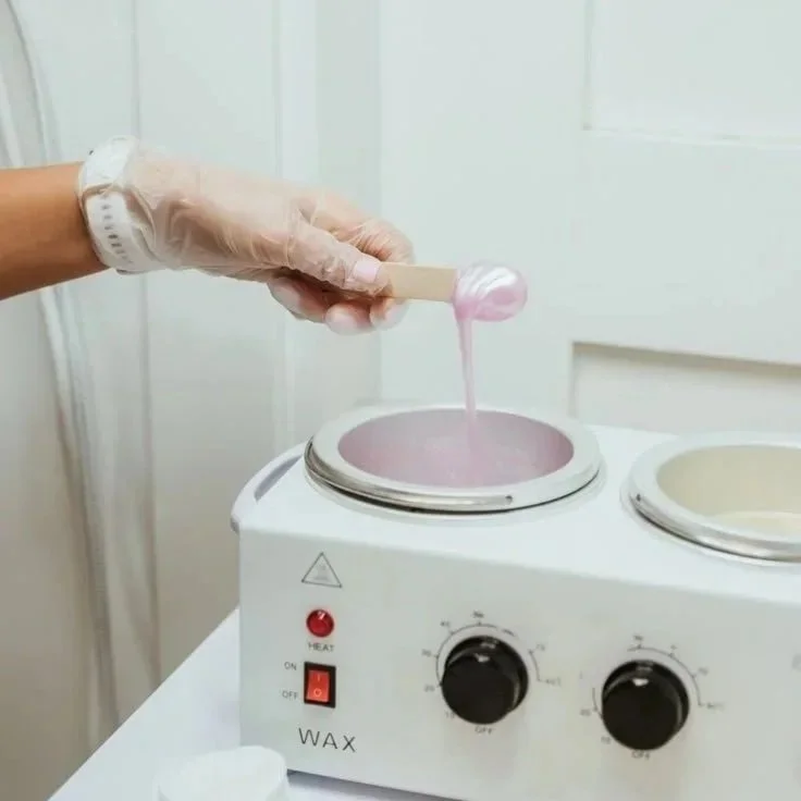 A person wearing gloves and a wristwatch is using a spatula to dip into pink wax in a small heating unit, likely for candle or cosmetic wax melting.
