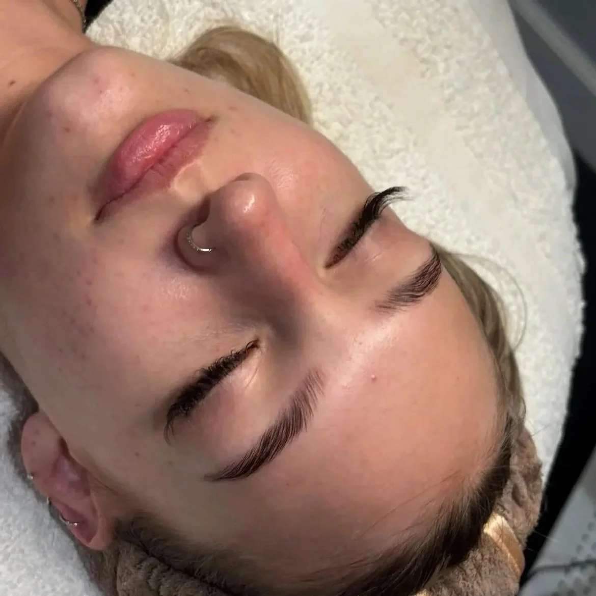 A close-up of a woman's face with closed eyes, lying on a white textured surface, showing her eyebrows, nose, lips, and facial skin.