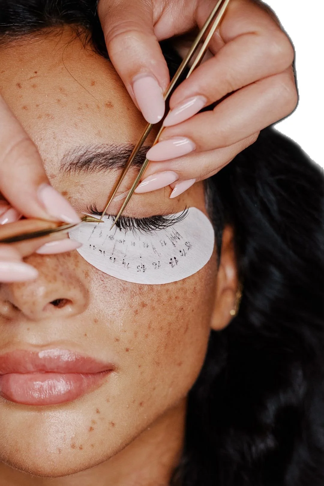A close-up of a woman getting her eyebrows waxed or shaped, with a white patch under her eyebrow and tweezers being used by a professional technician.