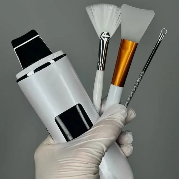 A hand wearing a latex glove holding a set of beauty tools, including a device, a fan brush, a spatula, and a small metal tool, against a gray background.
