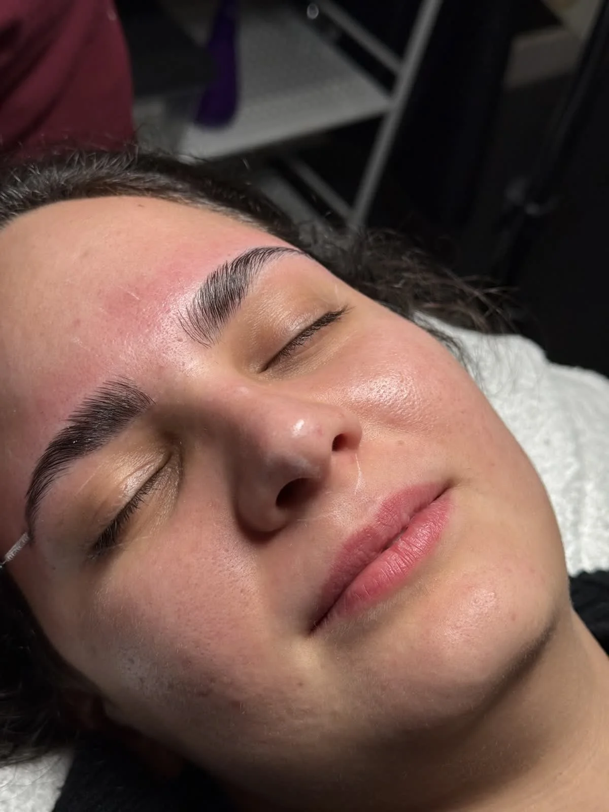 Close-up of a woman with her eyes closed, showing freshly done eyebrow microblading or similar cosmetic procedure, lying down with a relaxed expression.
