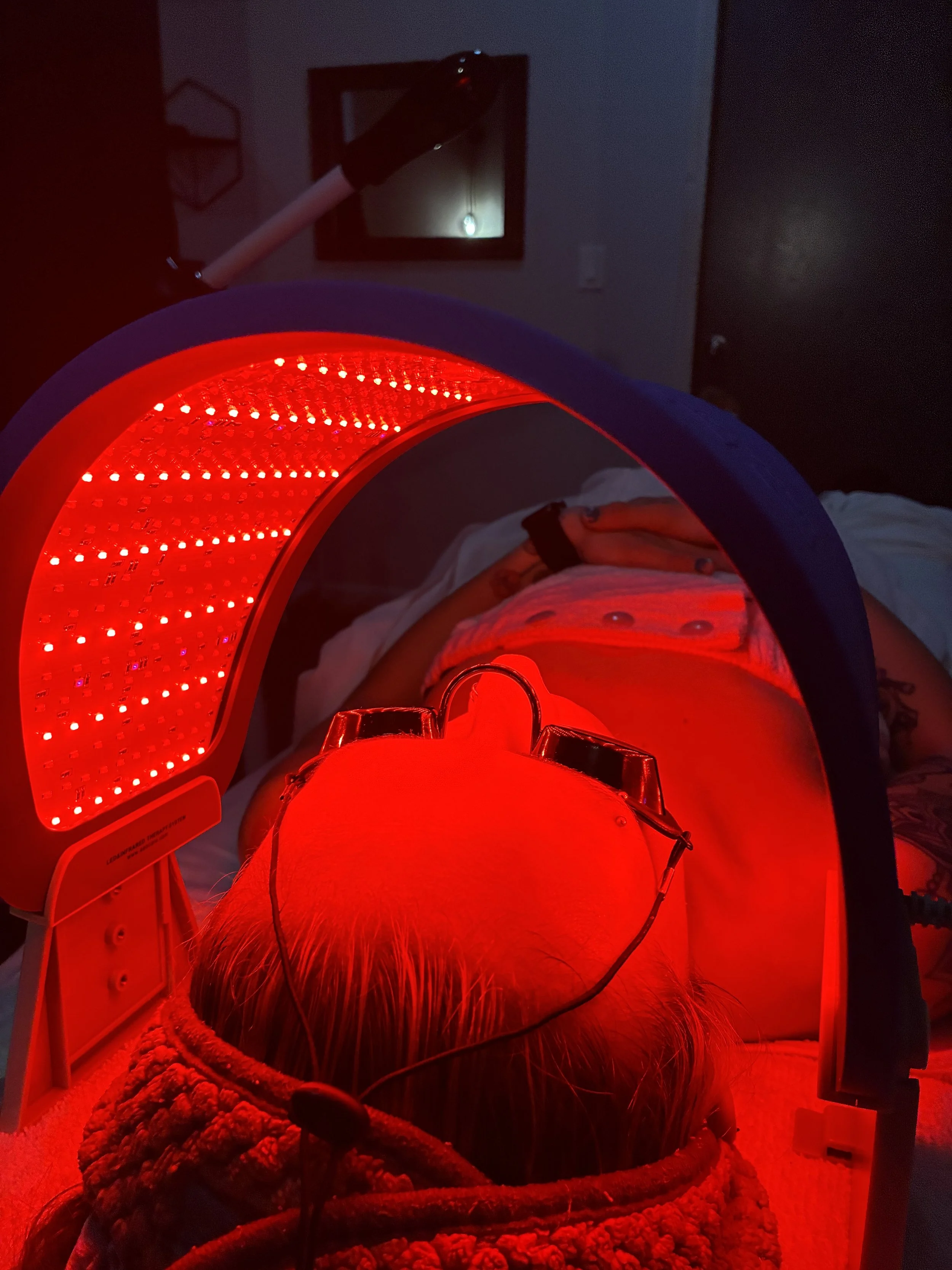 Person lying on a bed receiving red LED light therapy, with a red light panel overhead and a mirror on the wall reflecting a small light bulb in the background.