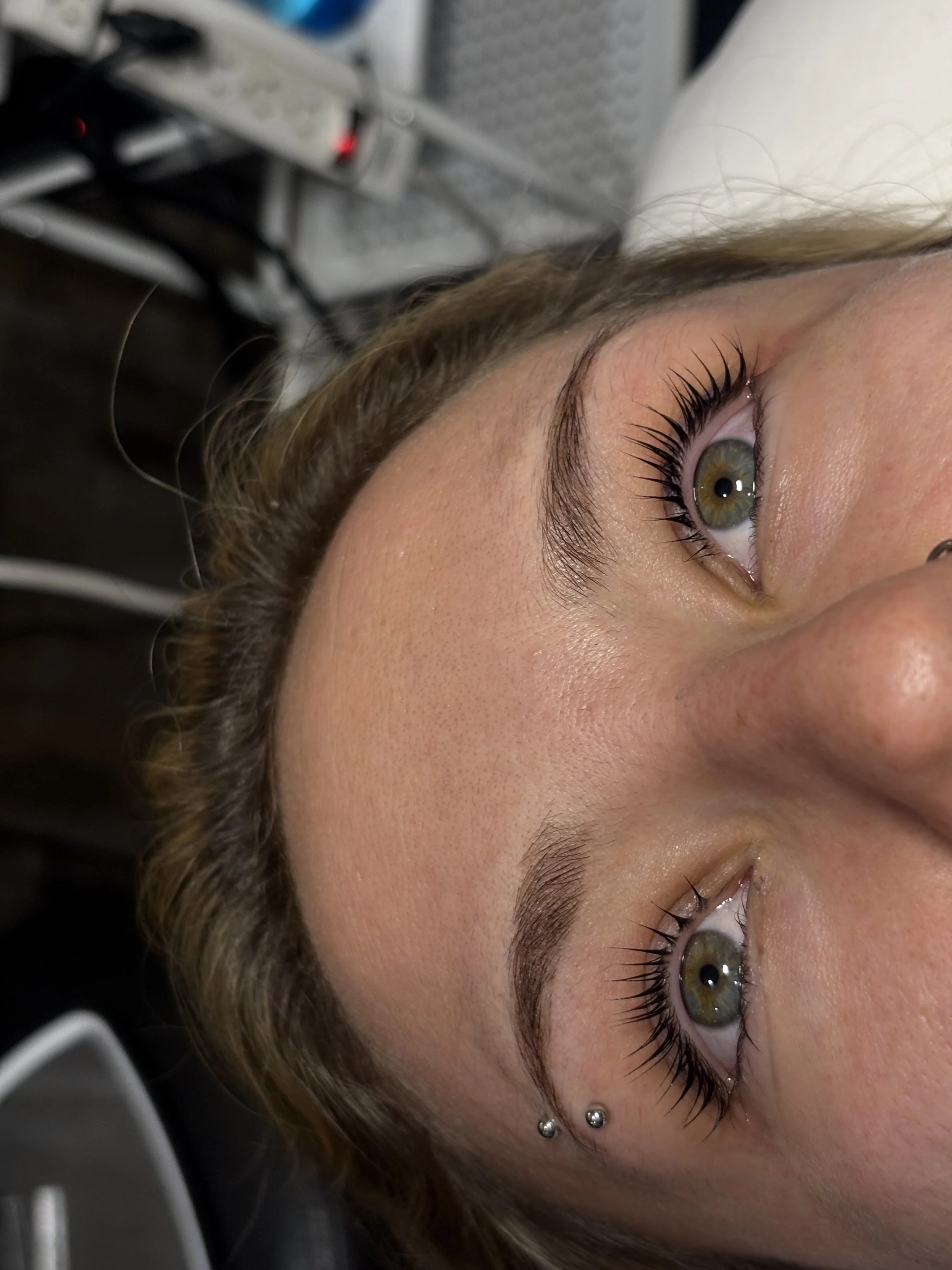 Close-up of a woman's face showing her green eyes, long eyelashes, and pierced ear with two silver studs, with a blurred background of medical equipment.