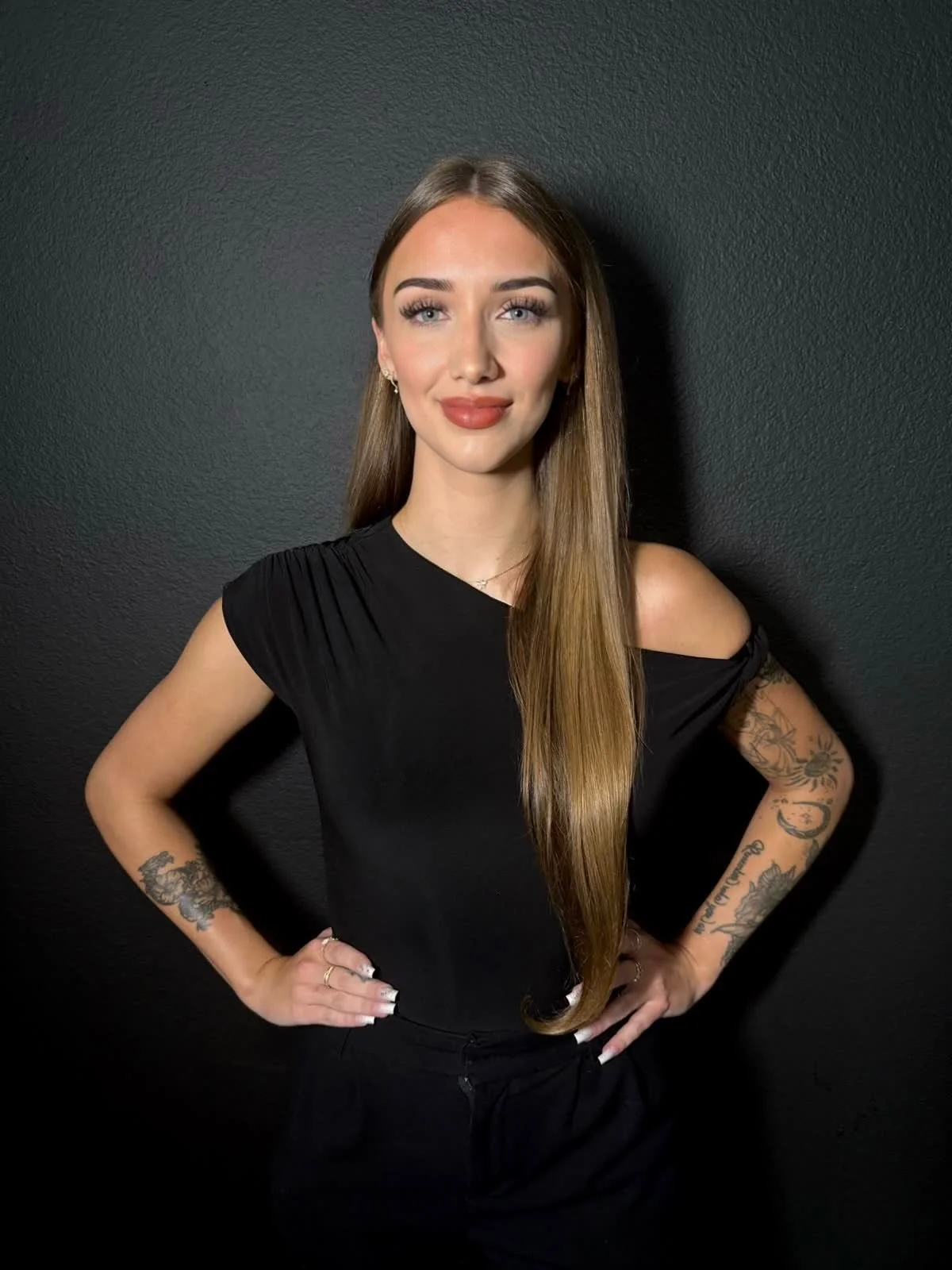 A woman with long, straight brown hair wearing a black top with shoulder cutouts and black pants, standing against a plain dark wall. She has tattoos on both arms, light makeup, and poses with her hands on her hips.
