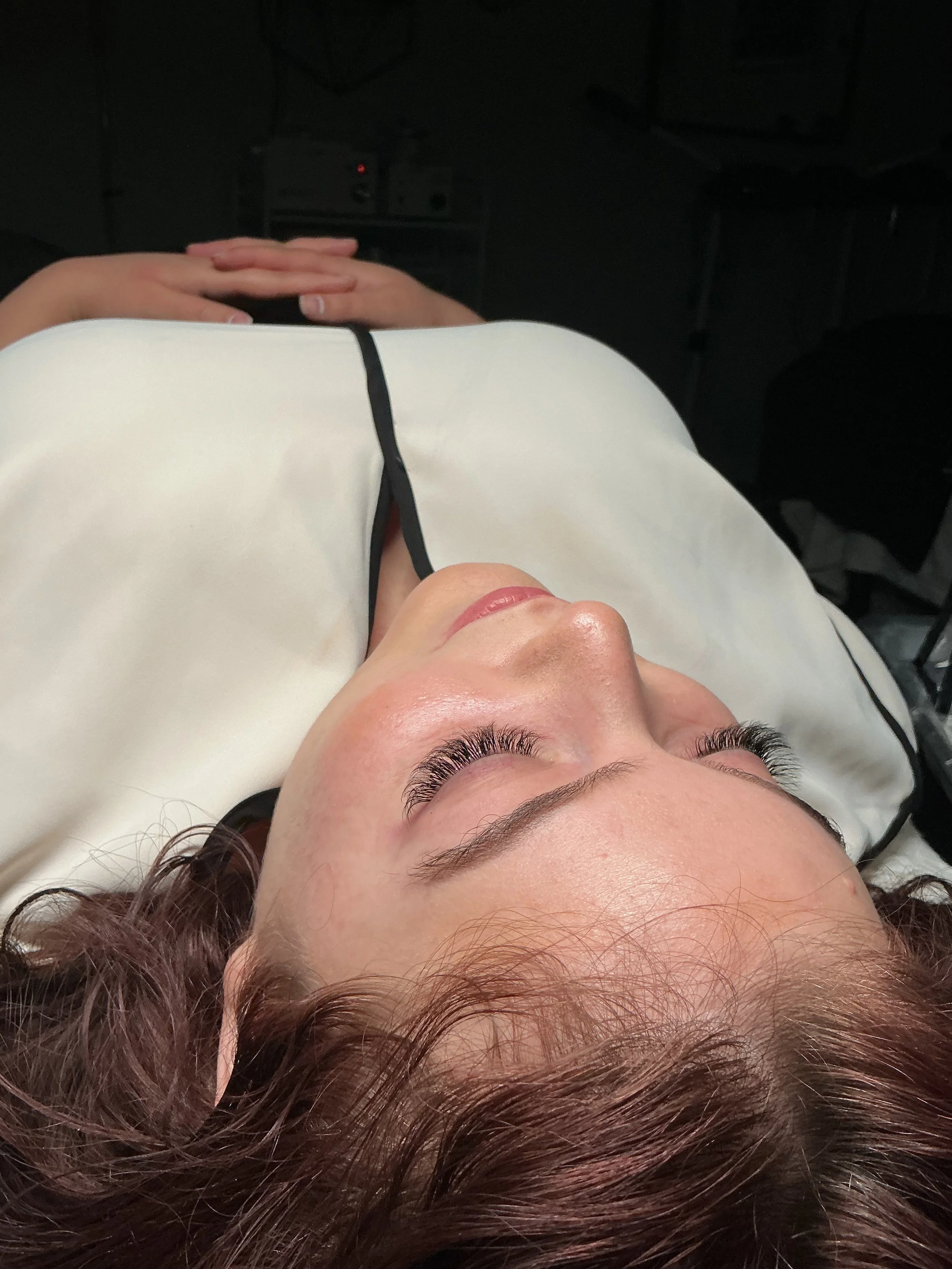 A person lying down with their eyes closed, wearing white clothing with black trim, and hands resting on their forehead, in a dark environment.