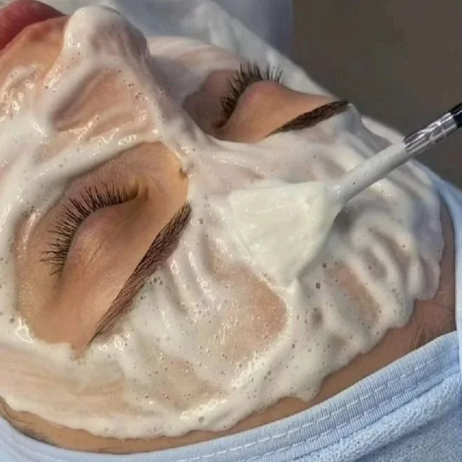 Close-up of a person's face covered in a foamy facial mask with closed eyes, receiving a facial treatment using a spatula.