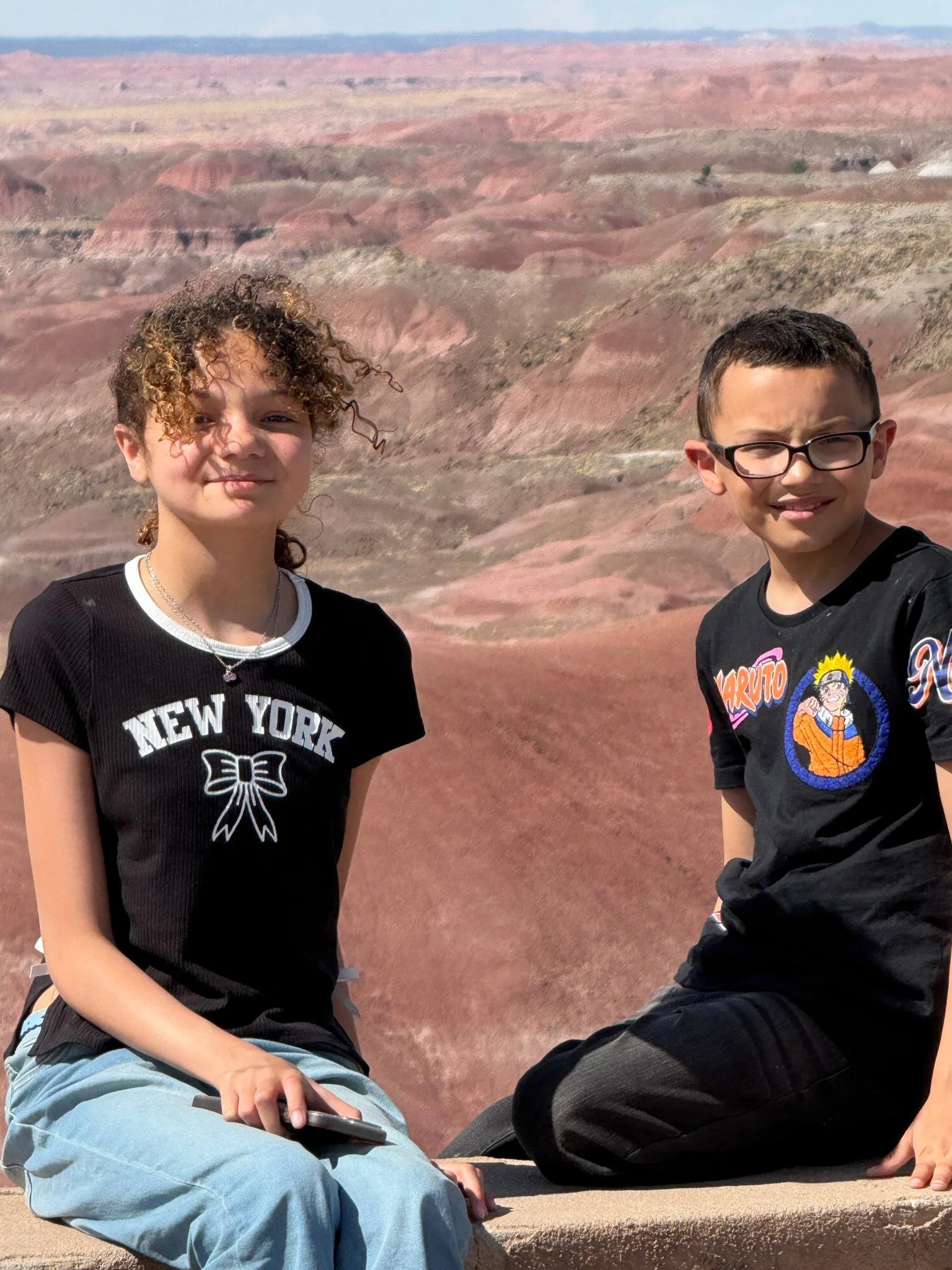 Two children sitting outdoors in front of colorful, layered canyon landscape under a clear sky.