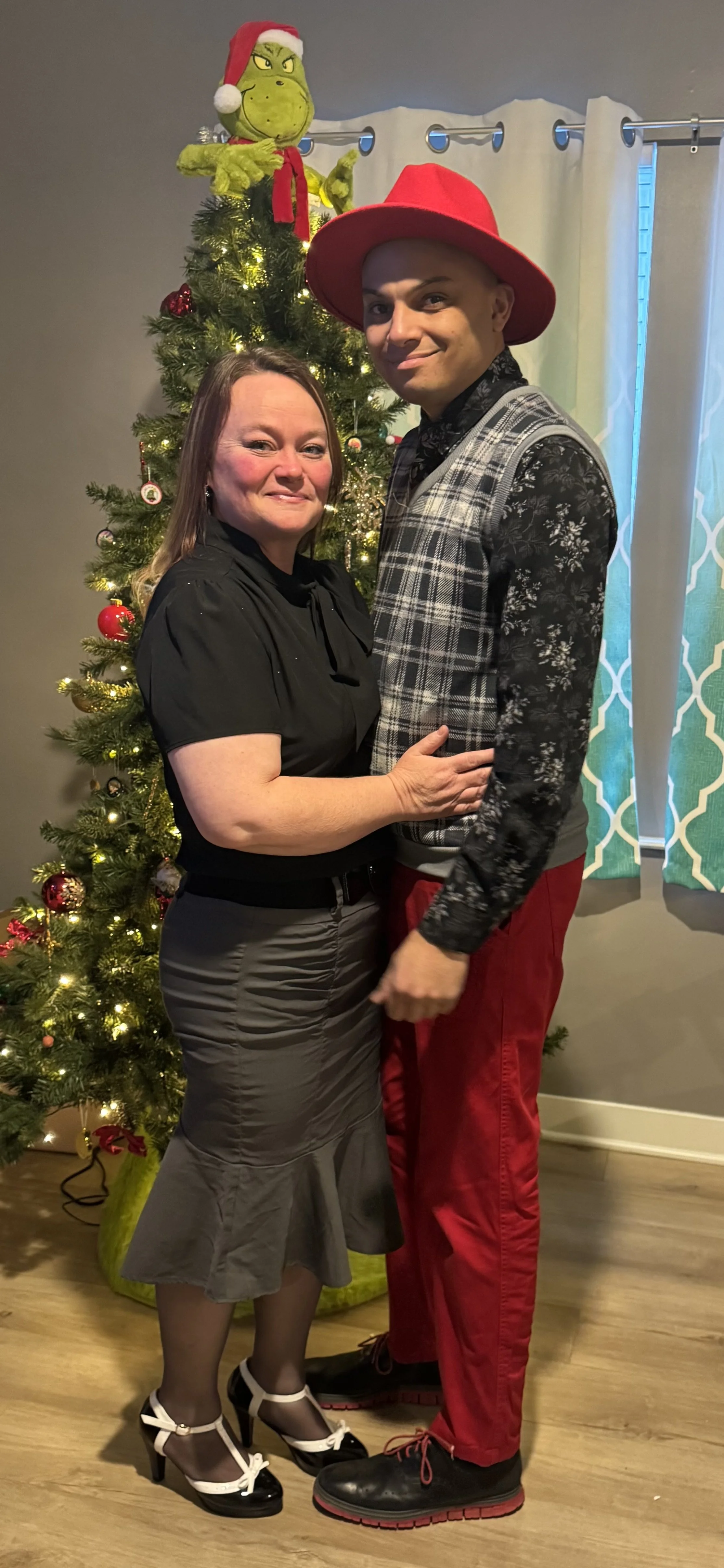 A woman and a man standing close together in front of a decorated Christmas tree, with a Grinch plush wearing a Santa hat on top, in a room with curtains and a window.