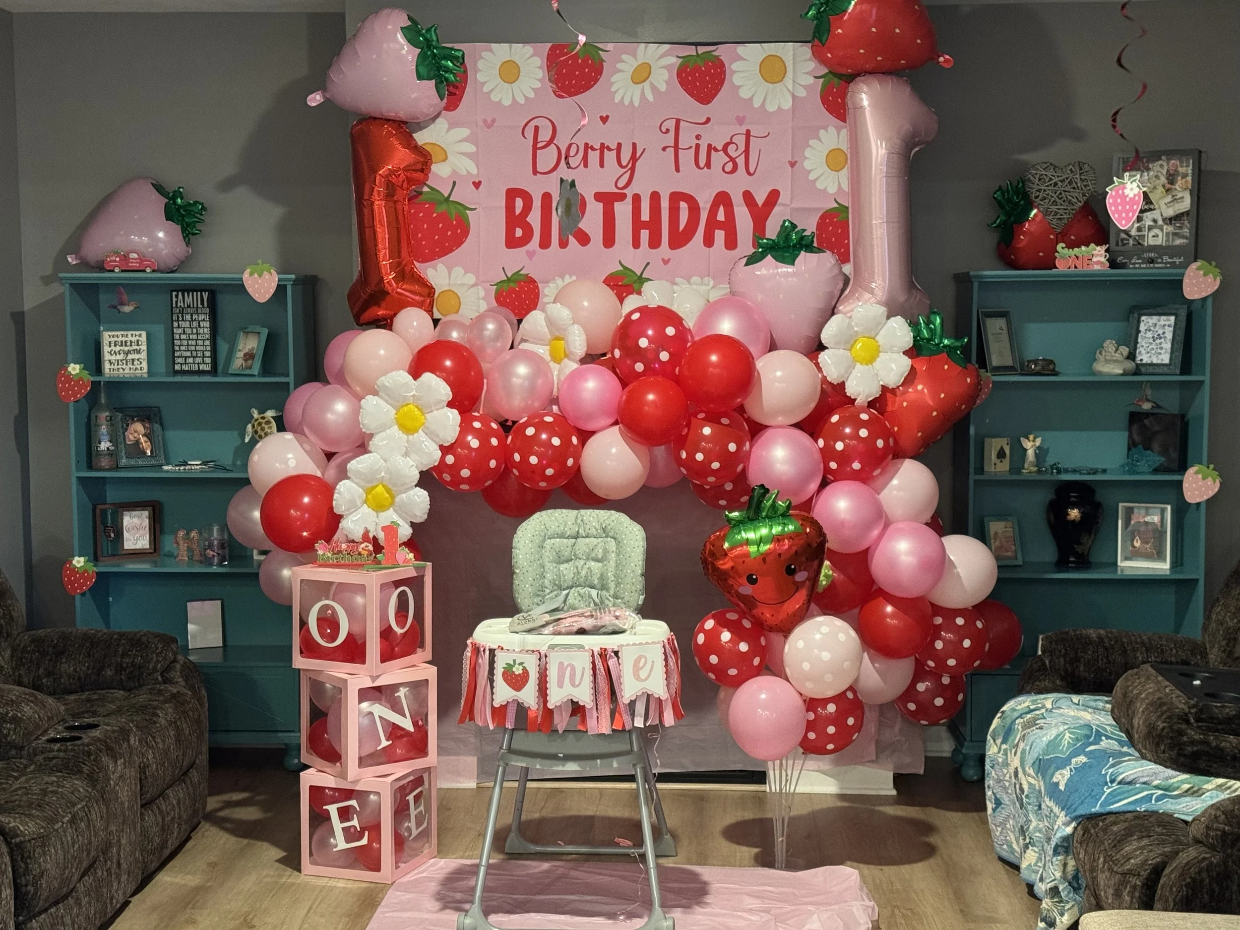 A birthday party setup with pink, red, and strawberry-themed balloons and decorations. A large pink banner reads 'Berry First Birthday'. There are shelves with framed photos and decorations, and a table with tableware and a pink birthday cake. The overall theme is strawberries and pinks.