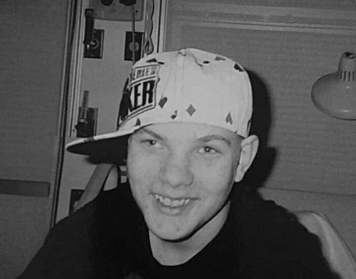 Smiling young man wearing a tilted cap with text, in a room with a lamp and electrical panel in the background.
