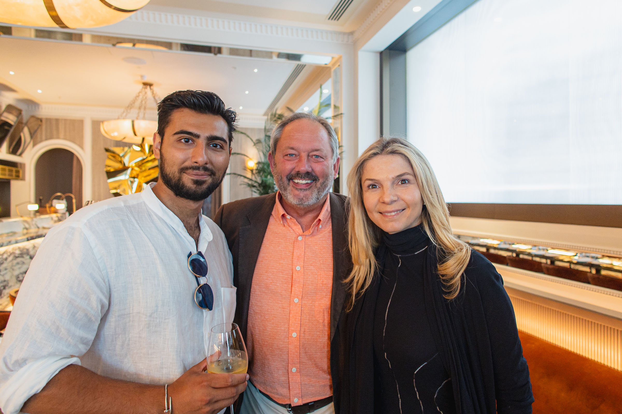 Three people posing together indoors at a social event, with a man holding a glass of wine.