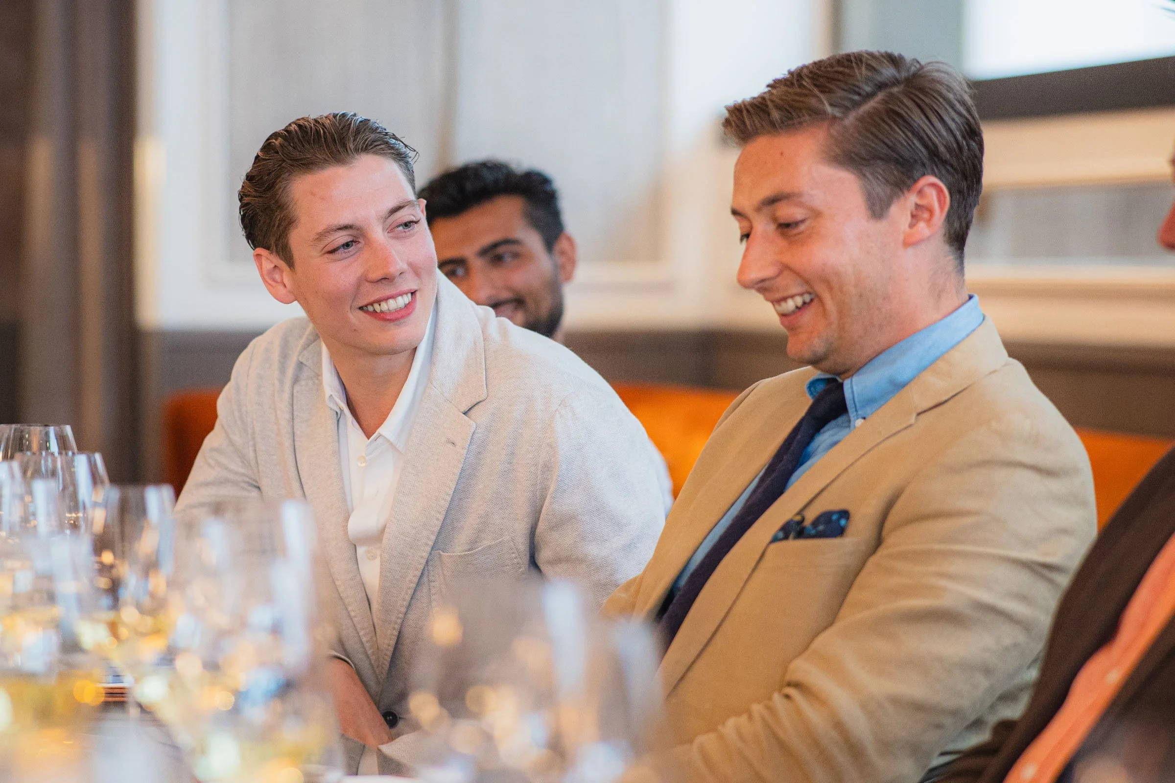 Three people, two men and one woman, sitting at a table in a restaurant, smiling and engaging in conversation. Glasses of water are visible on the table.