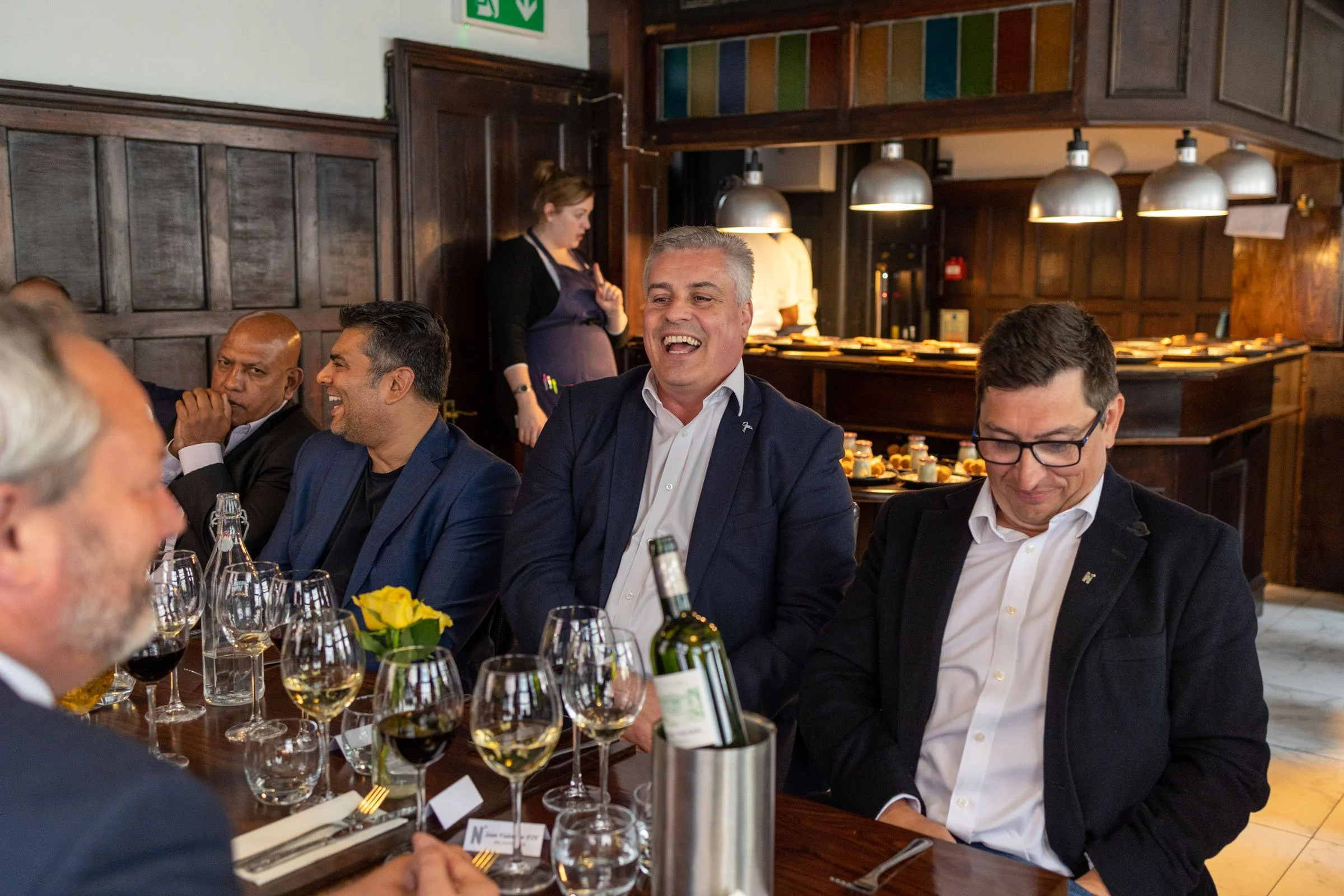 Group of men in business attire enjoying a meal and drinks in a restaurant, with one man smiling and laughing. The table is set with wine glasses, bottles, and a yellow flower centerpiece. A woman in an apron is standing in the background near a colorful glass display.