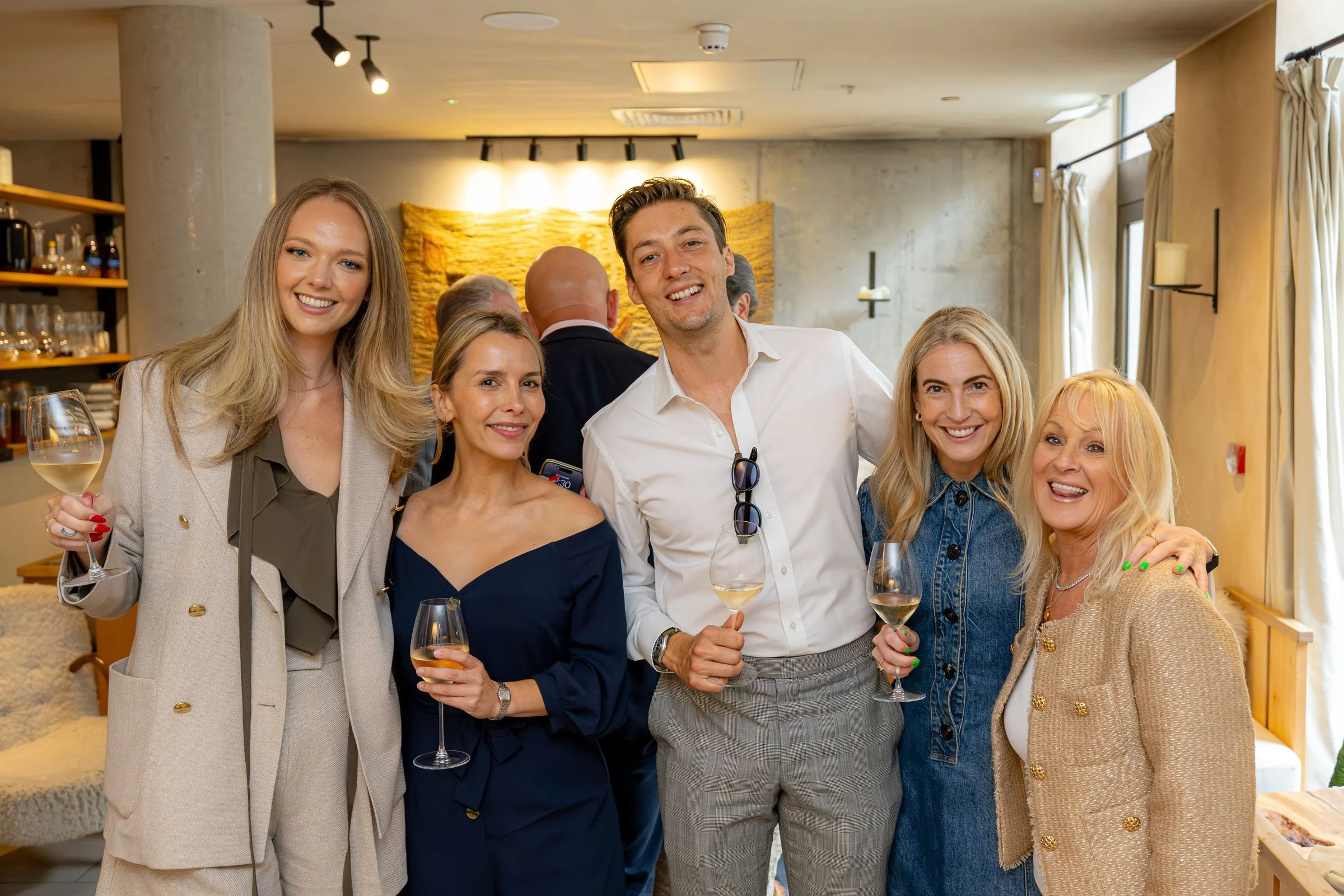 Group of six smiling adults in a restaurant, holding glasses of white wine, posing for a photo.