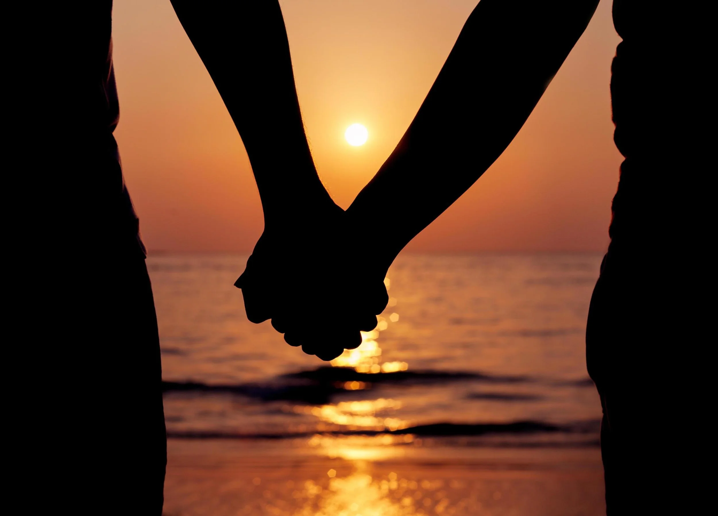 Silhouettes of two people holding hands at sunset by the water.