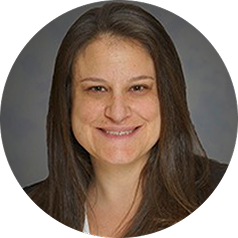A woman with long brown hair smiling at the camera, wearing a gray blazer.