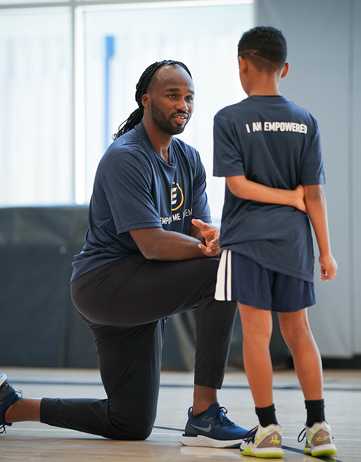 A man kneels on one knee and shakes hands with a young boy in a gym. Both are wearing navy blue t-shirts with the words 'I AM EMPOWERED' printed on the back and front, respectively. The man appears to be encouraging or coaching the boy.