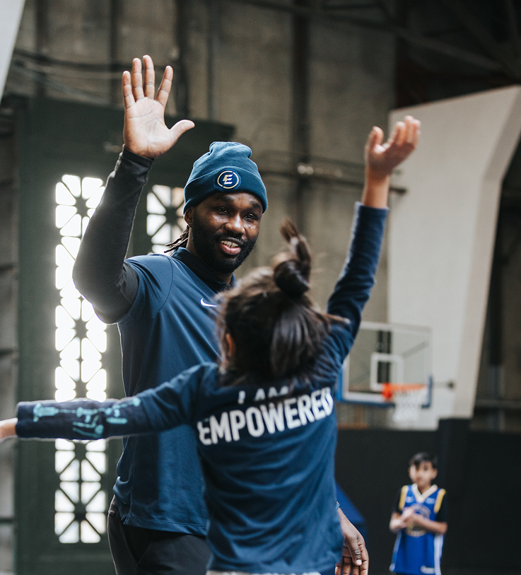 A man and young girl high-fiving inside a gymnasium, with a basketball hoop in the background. The girl is wearing a shirt with the word 'EMPOWERED' on the back.