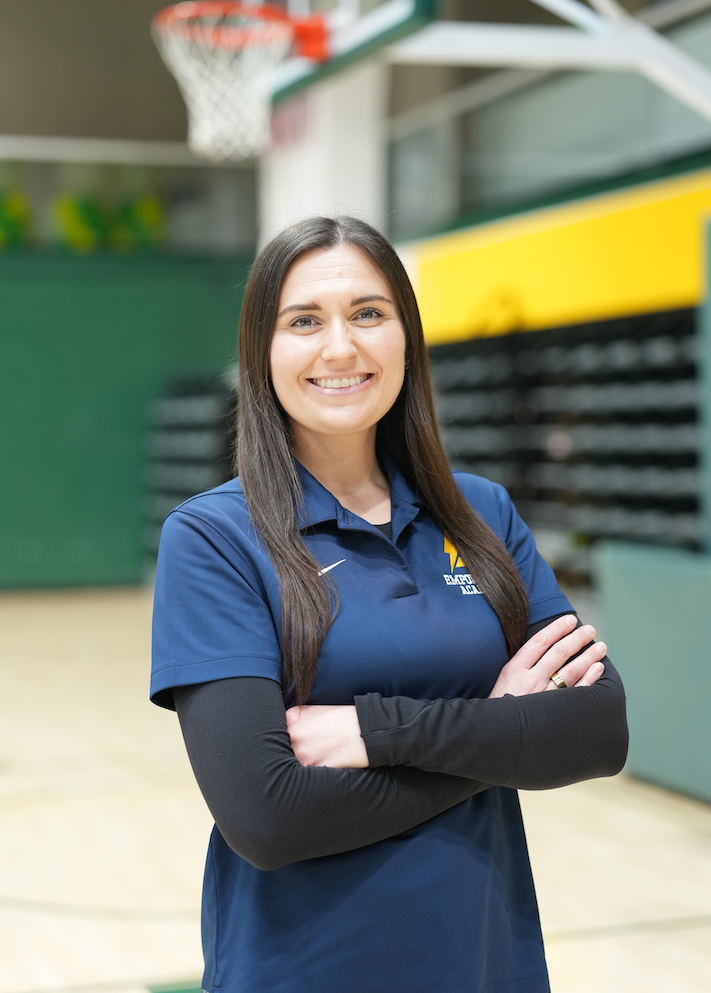 A woman with long brown hair smiling and crossing her arms, wearing a navy blue polo shirt with a logo, standing in an indoor gymnasium near basketball hoops.