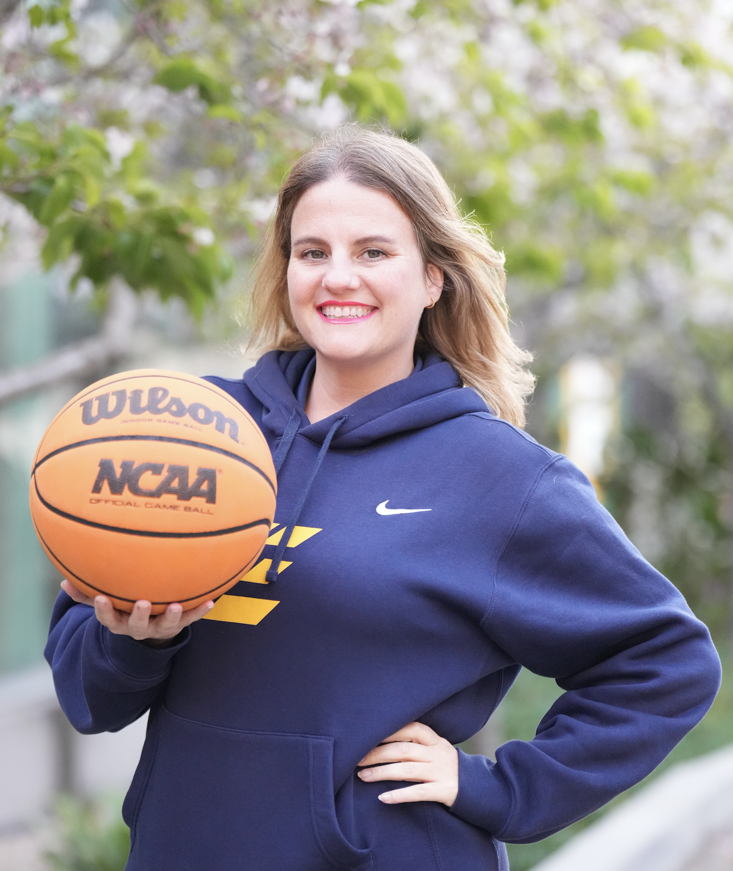 A woman smiling outdoors holding an NCAA official game basketball. She is wearing a navy blue hoodie with yellow and white graphics and has shoulder-length blonde hair. The background shows green trees.