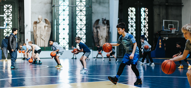 Children practicing basketball in an indoor gym with large windows and statues.