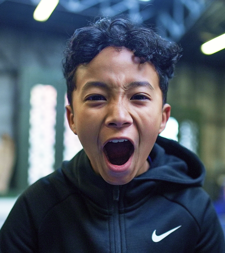 Young boy with curly hair and dark skin, wearing a black Nike jacket, is yelling or shouting with his mouth wide open indoors.