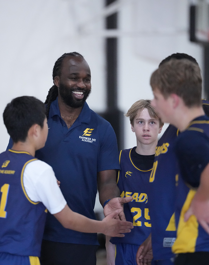 A basketball coach smiling and talking to young players in blue and yellow uniforms during a game or practice.