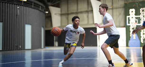 Two boys playing basketball on an indoor court, one dribbling the ball and the other guarding him.