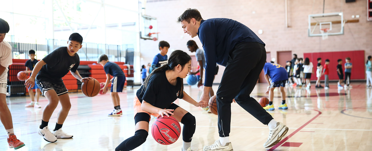 Coach instructs girl dribbling a basketball during practice with children in a gymnasium.