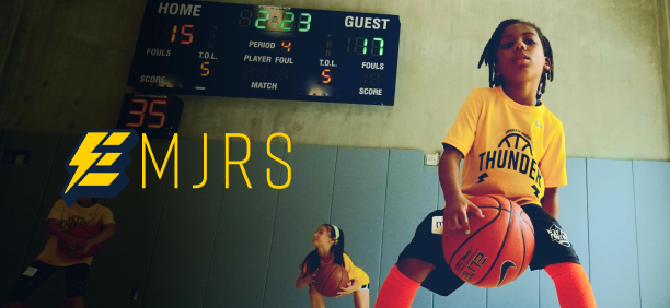 Young athlete holding a basketball during a drill in an indoor basketball court with a scoreboard overhead.