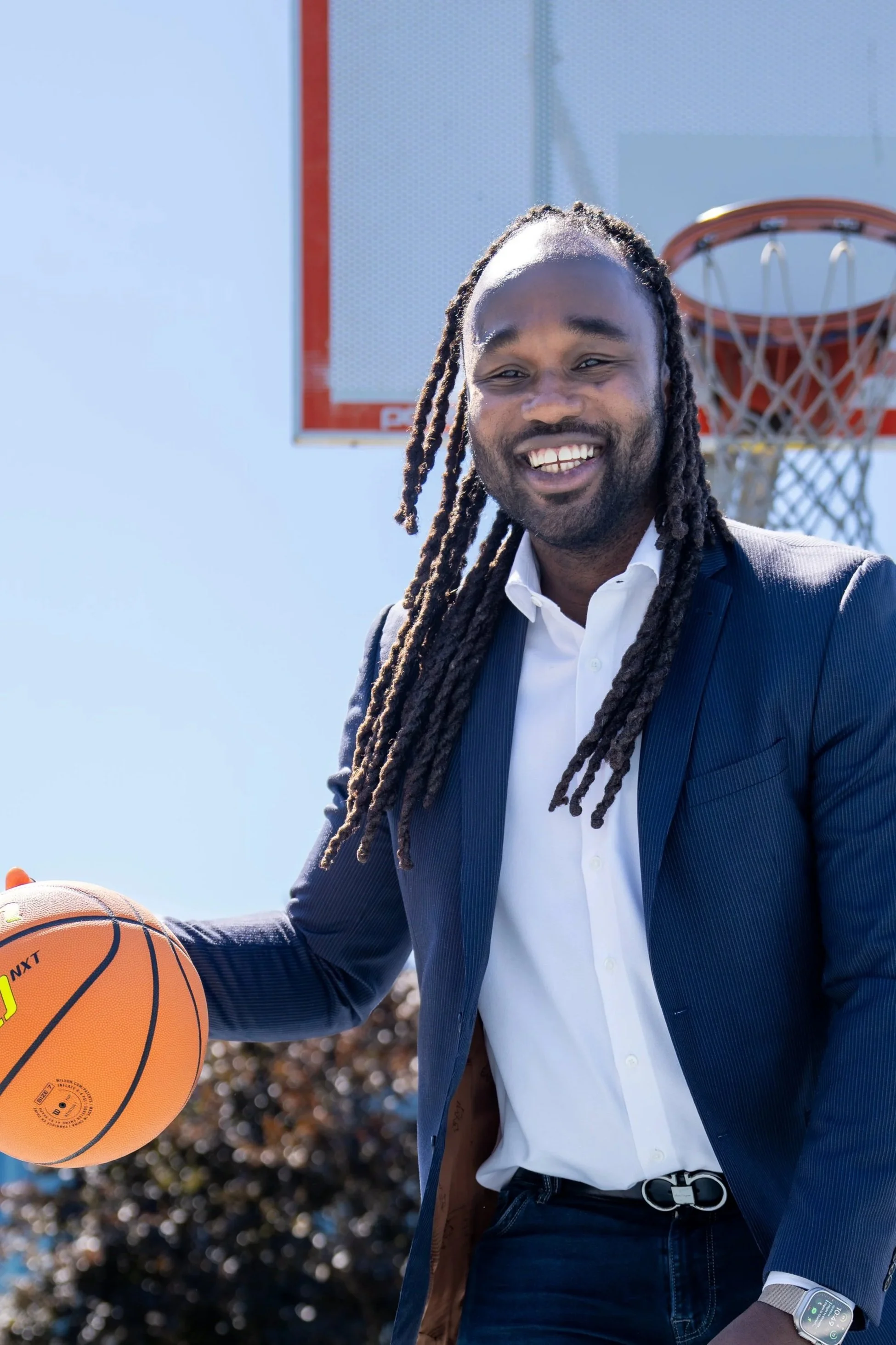 Coach Jerome Gumbs smiling and holding a basketball back with a basketball hoop in the background, outdoors on a sunny day.