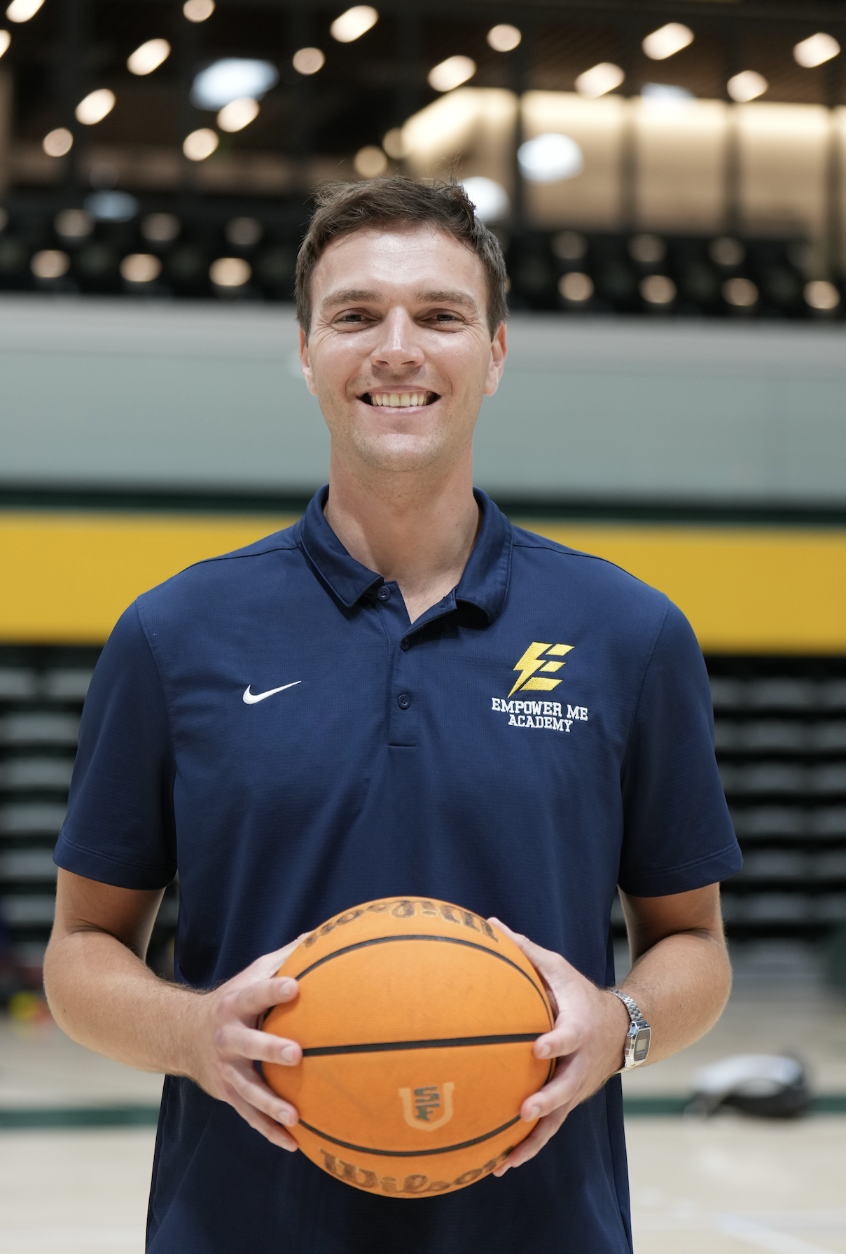 A smiling young man in a navy blue polo shirt, holding a basketball, standing inside a gymnasium with bleachers and bright lights.