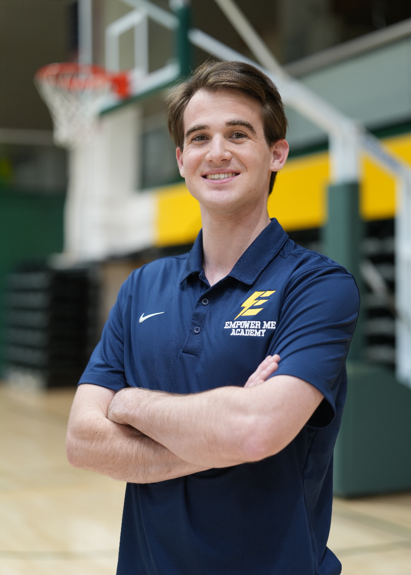 A young man with brown hair smiling, wearing a navy blue polo shirt with the "Empower Me Academy" logo, standing with arms crossed inside a gymnasium with a basketball hoop in the background.
