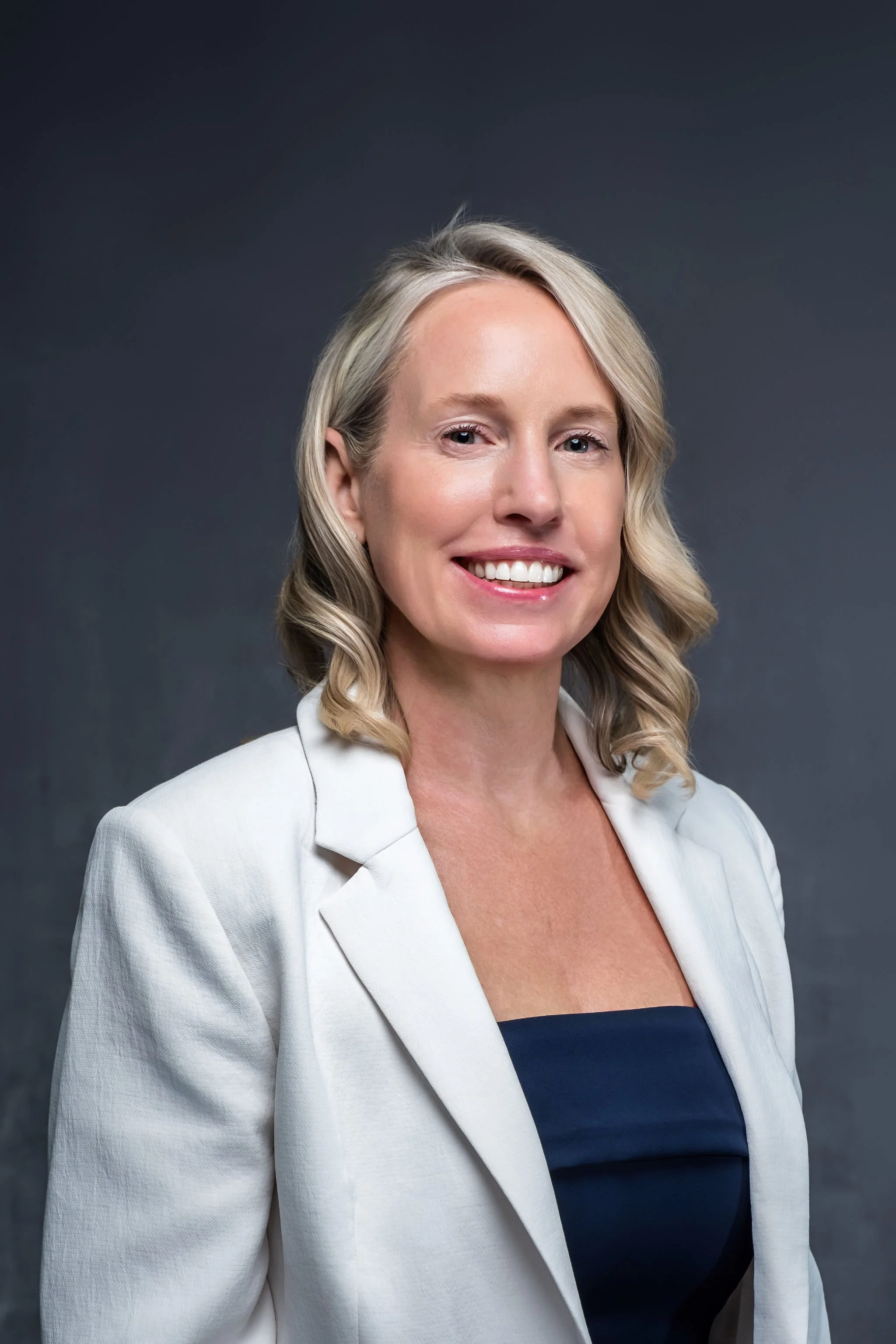 A woman with blonde hair styled in loose curls, smiling, wearing a white blazer over a dark top, against a dark gray background.