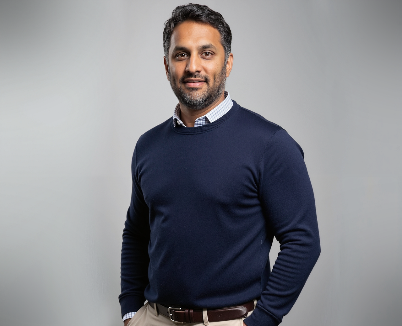 Professional man with dark hair, beard, wearing a navy sweater over a striped shirt, standing against a light gray background