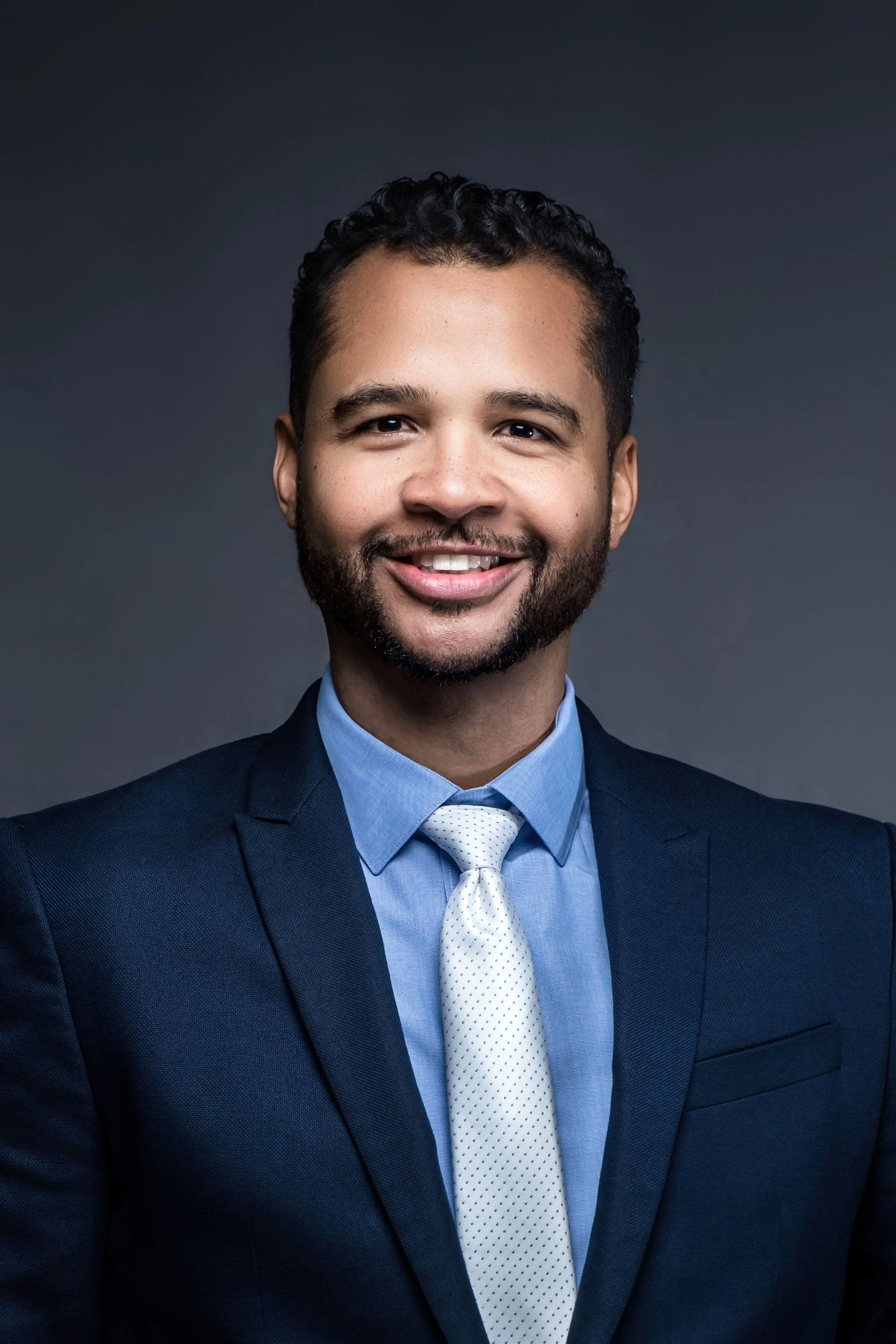 A smiling man in a blue suit, light blue shirt, and white tie, posing against a dark background.