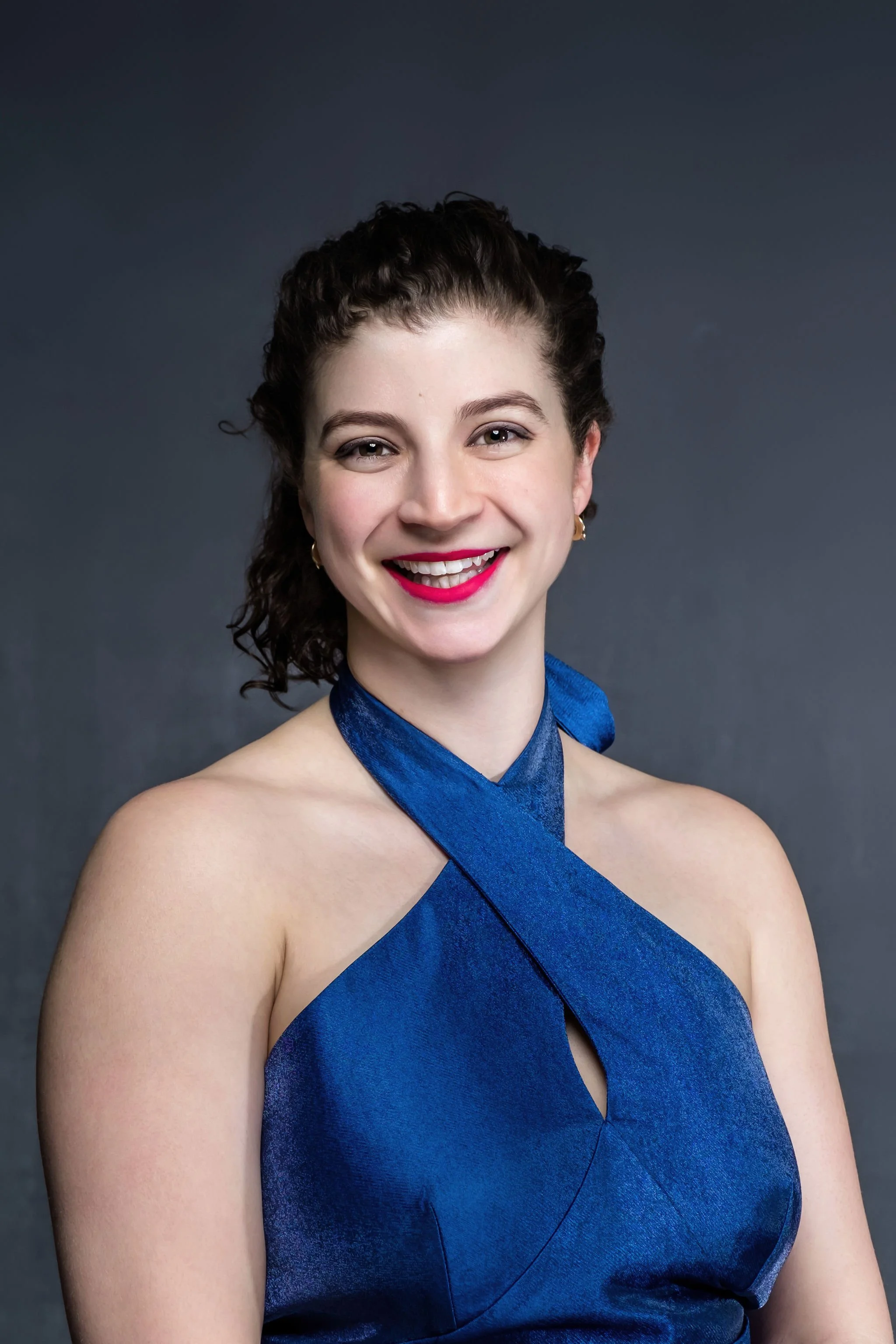 A woman with dark, curly hair smiling at the camera, wearing a blue halter dress and gold earrings, against a dark gray background.