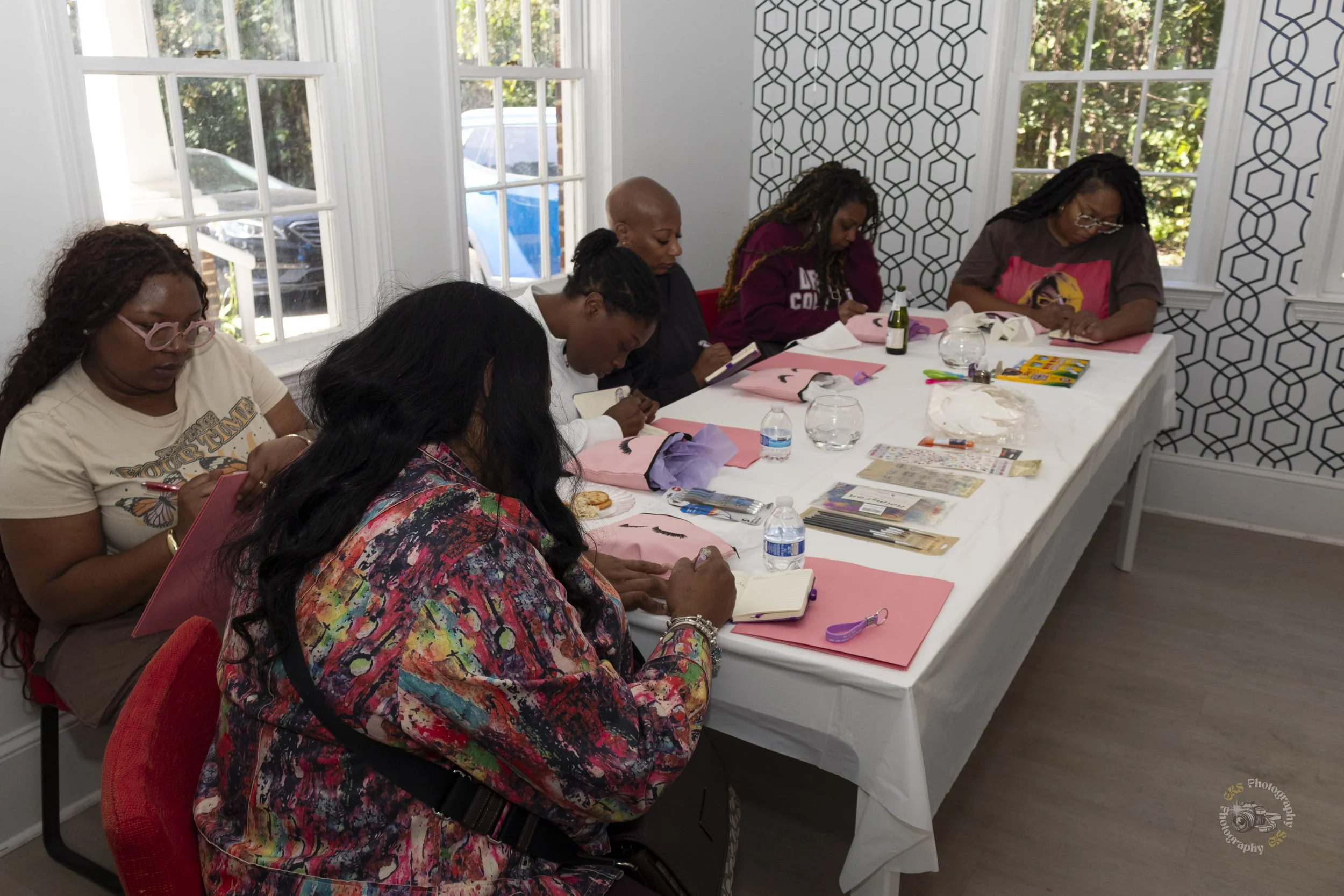 A group of six women sitting at a long table in a bright room, engaged in a creative activity. They are working on art projects with pink paper, makeup brushes, and various art supplies. The room has large windows with white frames, and a decorative patterned wall on one side.